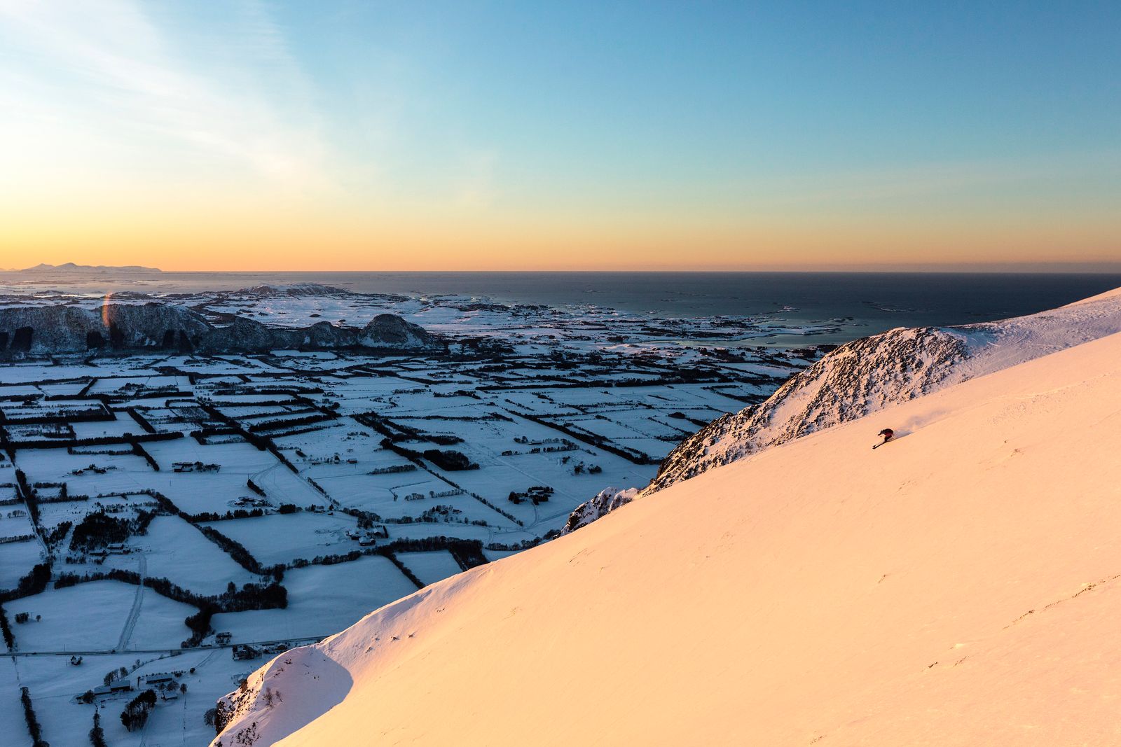 Skikjører på vei ned en bratt, snødekt fjellside i kveldssol, høyt over flatt kystlandskap og hav.